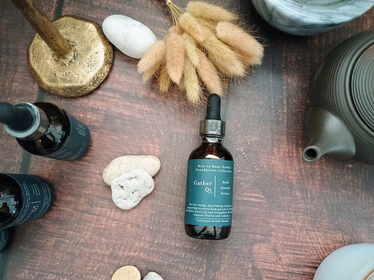 A small amber glass dropper bottle labeled 'Gather Qi' with a teal label and black dropper cap, placed on a dark wooden surface with stones, dried grass, and other bottles around it.