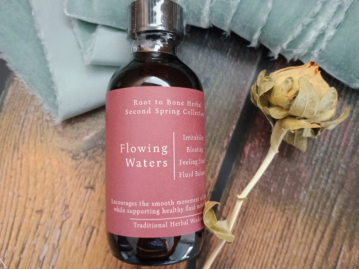 A bottle of Root to Bone Herbal 'Flowing Waters' tincture on a wooden surface next to a dried flower bud and a sage green cloth.