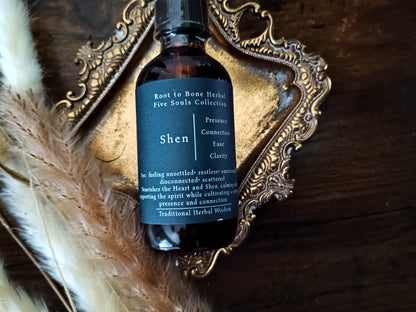 A dark amber glass bottle with a black label reading 'Shen' and 'Root to Bone Herbal Five Souls Collection', styled on an ornate gold tray over a dark wooden surface with dried grass and fur.