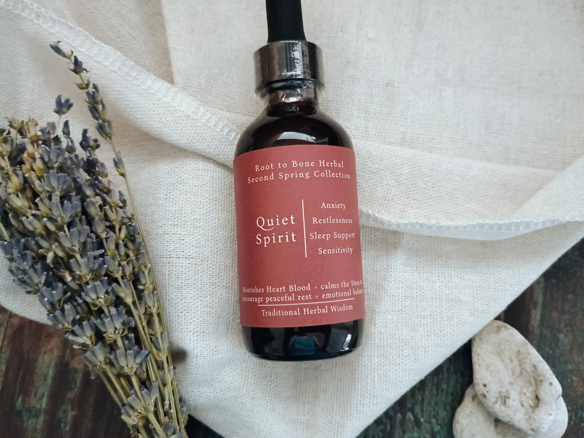A dark amber glass bottle of herbal sleep supplement with a black dropper cap and a dusty rose label reading 'Quiet Spirit'. The bottle is styled on a white linen cloth with dried lavender and small stones.