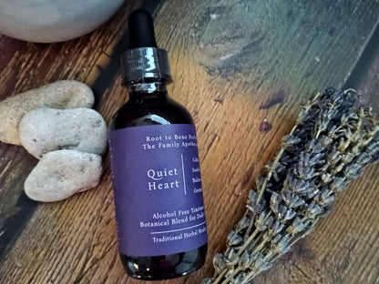 A dark amber glass dropper bottle labeled 'Quiet Heart' by Root to Bone Herbs, styled on a rustic wooden surface with smooth stones and a dried lavender sprig.