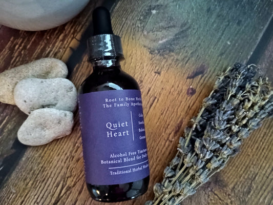 A dark amber glass dropper bottle labeled 'Quiet Heart' by Root to Bone Herbs, styled on a rustic wooden surface with smooth stones and a dried lavender sprig.