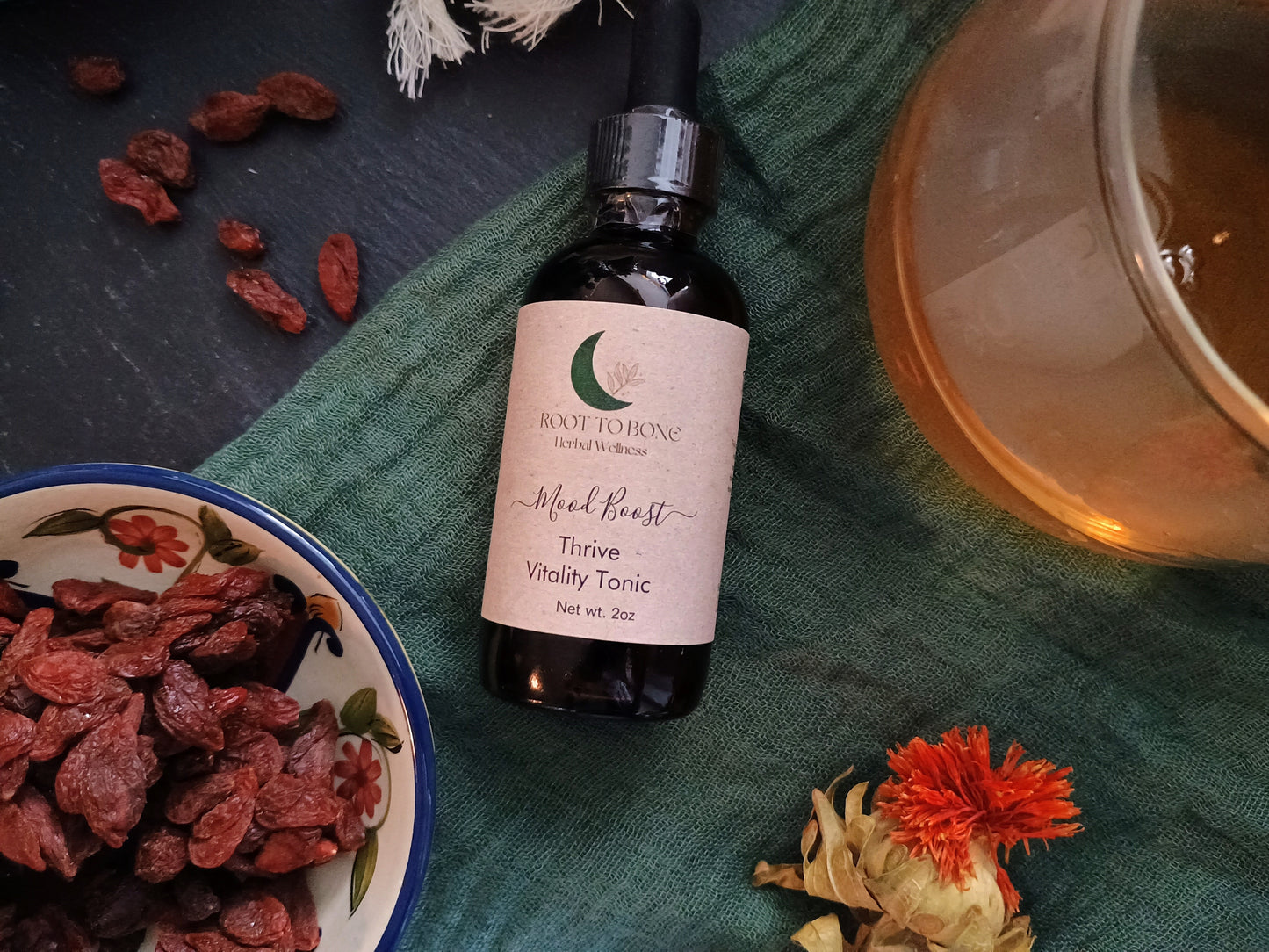 Bottle of 'Mood Boost' Vitality Tonic with dried berries and a cup on a green cloth.