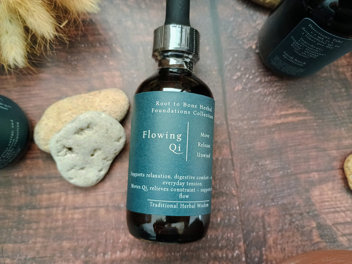 A dark amber glass dropper bottle labeled 'Flowing Qi' by Root to Bone Herbal, placed on a rustic wooden surface with decorative stones and dried grass.