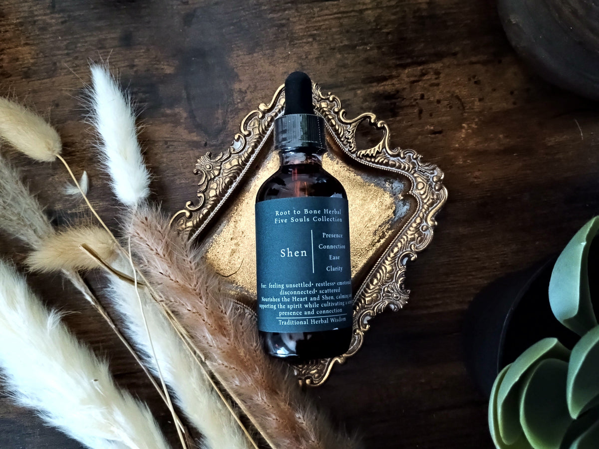 A dark amber glass dropper bottle labeled 'Shen' by Root to Bone Herbs, styled on an ornate gold tray with dried pampas grass and a succulent on a dark wooden surface.