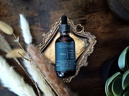 A dark amber glass dropper bottle labeled 'Shen' by Root to Bone Herbs, styled on an ornate gold tray with dried pampas grass and a succulent on a dark wooden surface.