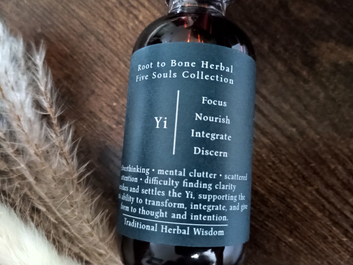 A dark amber glass bottle of Root to Bone Herbal Five Souls Collection 'Yi' tincture, featuring a slate blue label, styled on a wooden surface next to dried feathers.