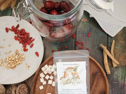 A clear stand-up pouch labeled 'Nourishing Earth Blend' sits on a wooden tray beside loose herbal ingredients, including red goji berries, beige seeds, white cubes, and brown roots, with a glass jar of red dried fruit pieces on a rustic wooden surface.