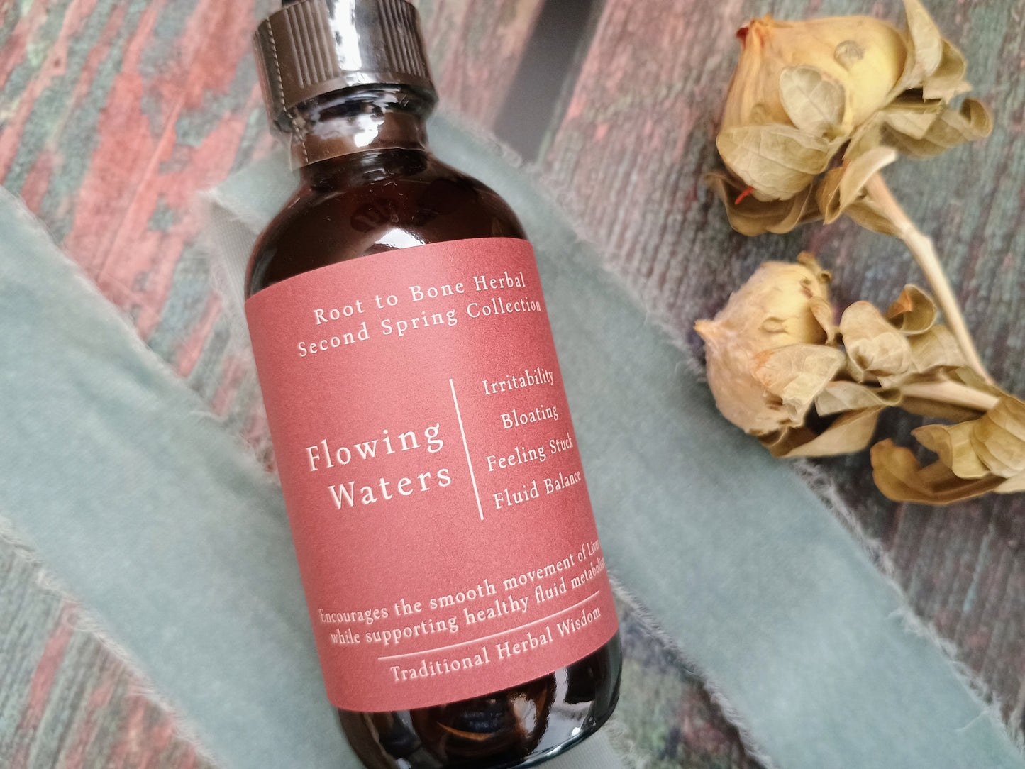A small amber glass bottle with a dark pink label reading 'Root to Bone Herbal Second Spring Collection' and 'Flowing Waters,' styled on a textured surface with dried flowers and a pale blue cloth.