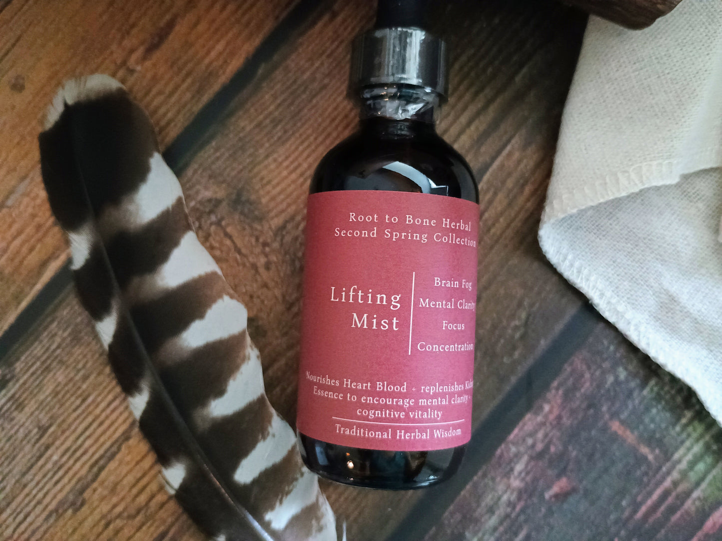 A dark amber glass dropper bottle labeled 'Lifting Mist' by Root to Bone Herbal, resting on a rustic wooden surface next to a black and white tiger stripe feather and a folded beige cloth.