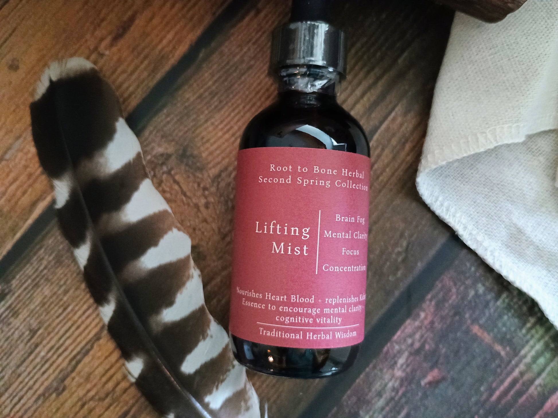 A dark amber glass dropper bottle labeled 'Lifting Mist' by Root to Bone Herbal, resting on a rustic wooden surface next to a black and white tiger stripe feather and a folded beige cloth.