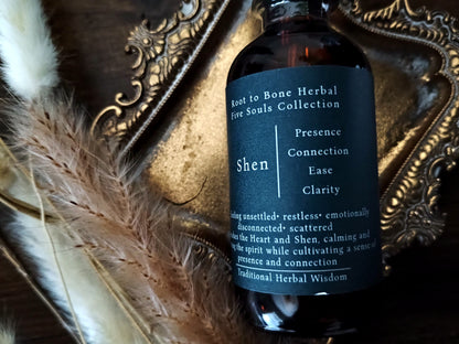 An amber glass bottle of Root to Bone Herbal Five Souls Collection, labeled 'Shen', styled on a dark wood surface with a gold ornate tray and dried pampas grass.