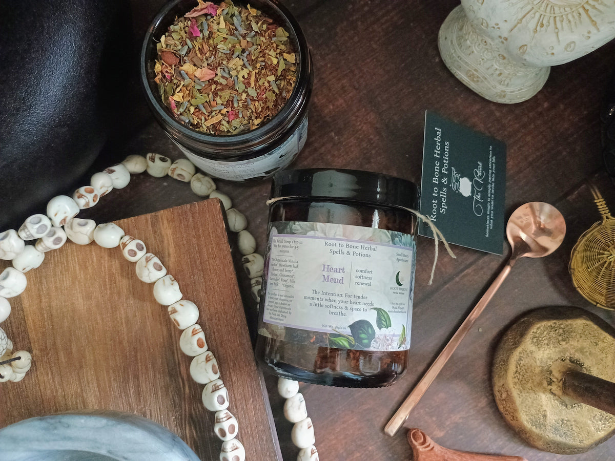 A styled flat-lay of a jar of herbal tea labeled 'Heart Mend' on a dark wooden surface, surrounded by decorative props including a skull bead necklace, a copper spoon, a mortar and pestle, and a dried flower jar.