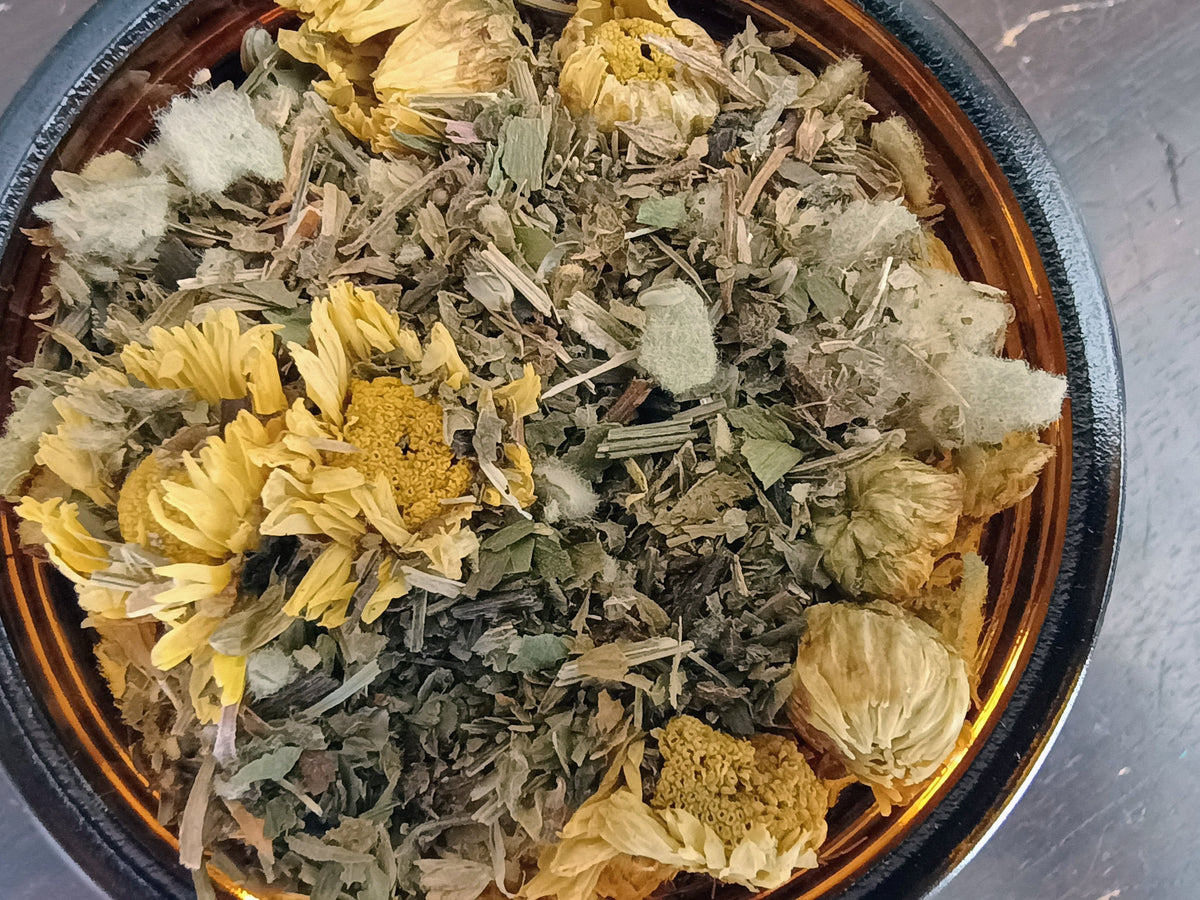 Dried herbal tea blend with yellow chamomile flowers and green leaves in an amber glass bowl.