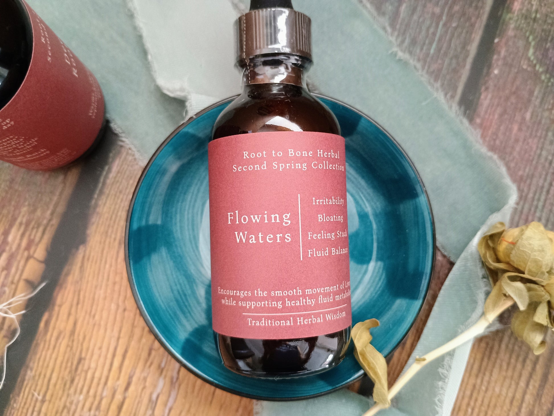 A dark amber glass bottle of herbal tincture with a pink label reading 'Flowing Waters' and 'Root to Bone Herbal Collection,' placed on a teal ceramic dish over a wooden surface with dried flowers and a second bottle partially visible.