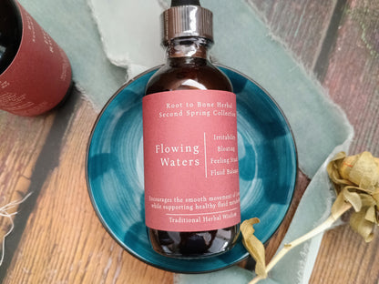 A dark amber glass bottle of herbal tincture with a pink label reading 'Flowing Waters' and 'Root to Bone Herbal Collection,' placed on a teal ceramic dish over a wooden surface with dried flowers and a second bottle partially visible.