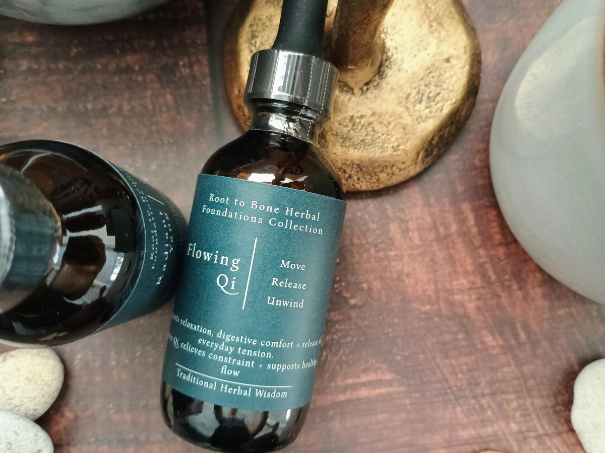A dark amber glass dropper bottle labeled 'Flowing Qi' from the Root to Bone Herbal Foundations Collection, styled on a rustic wooden surface with a gold-toned stone and white stones nearby.