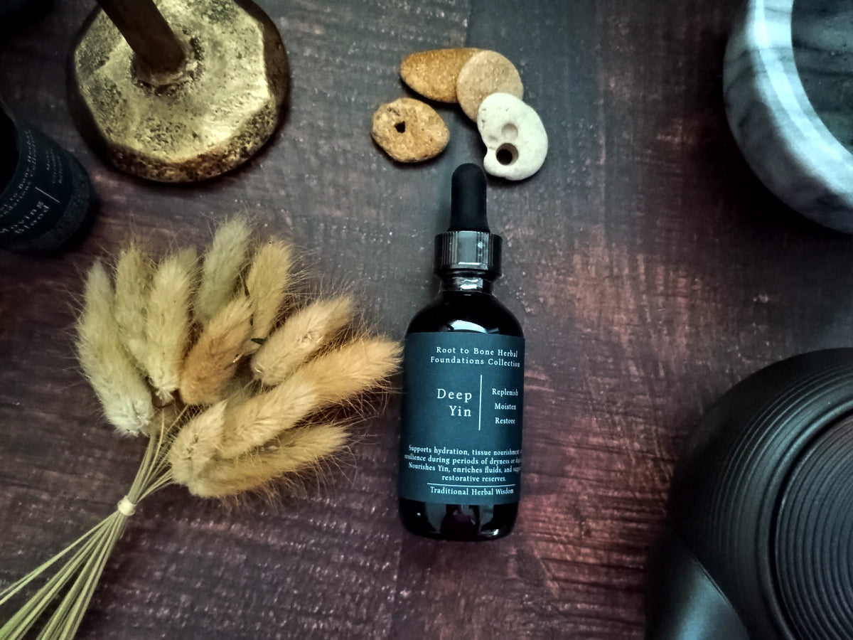 A dark amber glass dropper bottle labeled 'Deep Yin' by Root to Bone Herbals, styled on a dark wooden surface with dried bunny tail grass, small stones, and a brass candle holder.