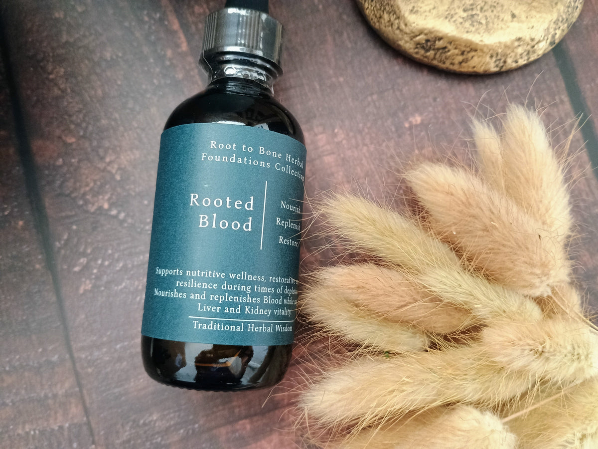 A dark amber glass dropper bottle labeled 'Rooted Blood' by Root to Bone Herbs, placed on a dark textured surface next to dried beige bunny tails and a stone.