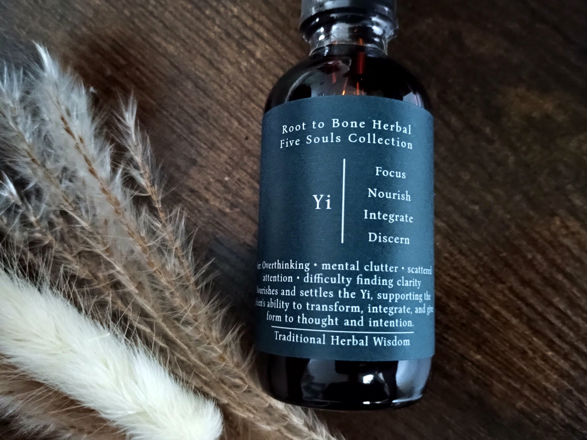 A dark amber glass bottle with a black cap and a dark teal label reading 'Root to Bone Herbal Five Souls Collection Yi', placed on a wooden surface next to dried white and brown grasses.