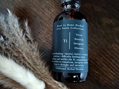 A dark amber glass bottle with a black cap and a dark teal label reading 'Root to Bone Herbal Five Souls Collection Yi', placed on a wooden surface next to dried white and brown grasses.