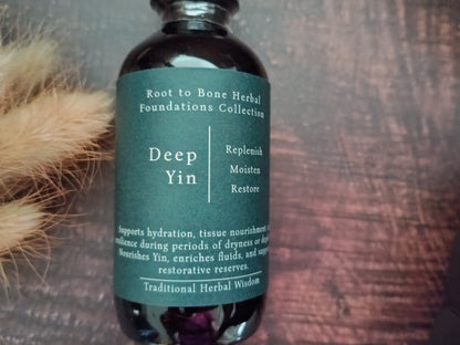 A dark amber glass bottle with a dark green label reading 'Root to Bone Herbal Foundations Collection Deep Yin' on a rustic wooden background with dried grass on the left.