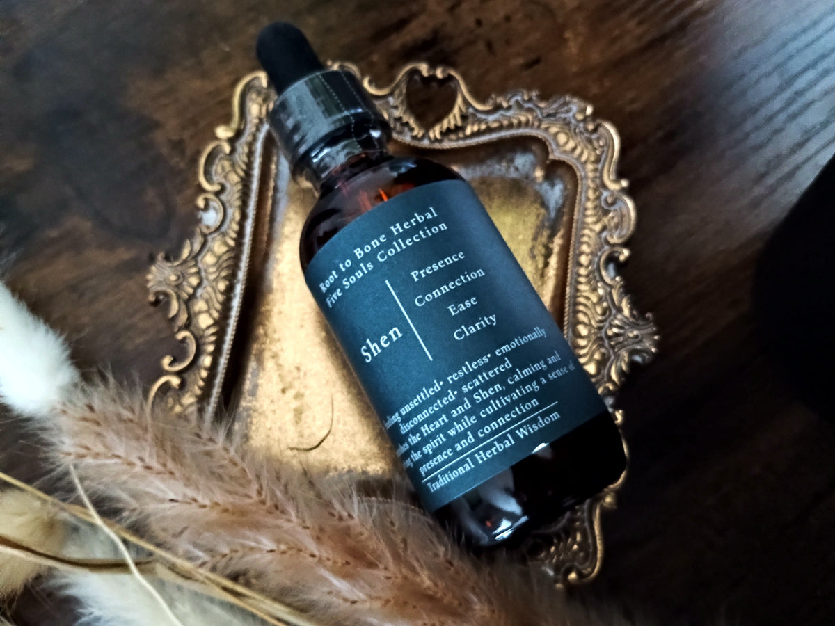 A dark amber glass dropper bottle with a black label reading 'Shen' and 'Root to Bone Herbal Apothecary Collection,' styled on an ornate gold tray over a dark wooden surface with dried grass and soft white fibers.