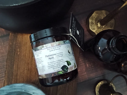 A dark amber glass jar candle labeled 'Protection Spell' by Root to bone, styled on a dark wooden surface with occult-themed props including a brass athame, a black apothecary bottle, and a black bowl.