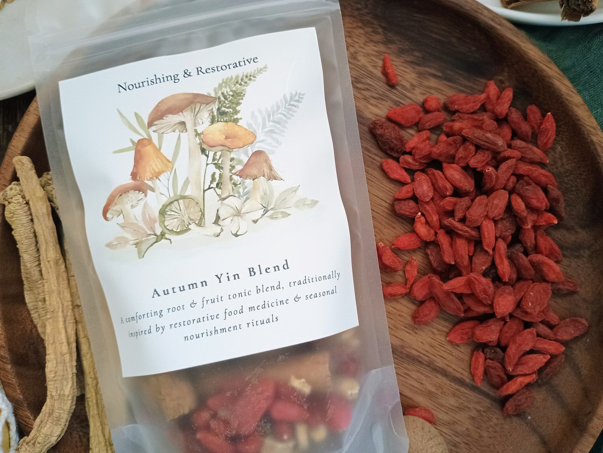 A clear stand-up pouch labeled 'Autumn Yin Blend' with mushroom and botanical artwork, placed on a wooden tray beside loose red goji berries and dried roots. The product appears to be a herbal tea or tonic blend.