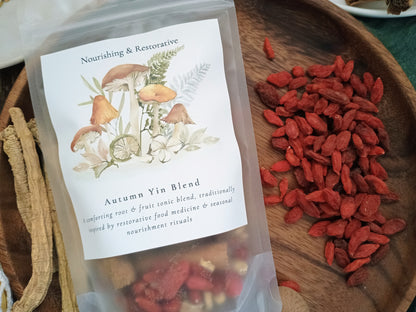 A clear stand-up pouch labeled 'Autumn Yin Blend' with mushroom and botanical artwork, placed on a wooden tray beside loose red goji berries and dried roots. The product appears to be a herbal tea or tonic blend.