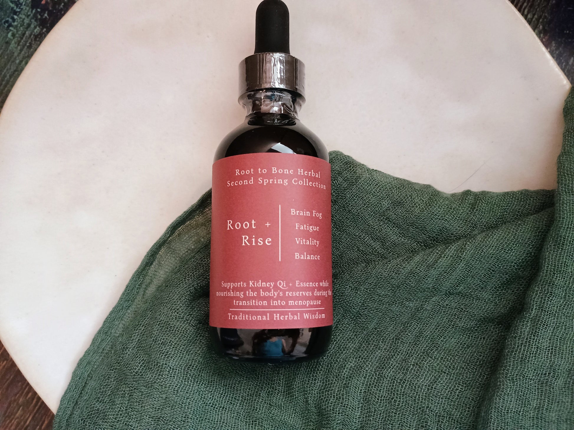 A glass dropper bottle of Root + Rise herbal tincture with a dusty rose label, resting on a textured green cloth over a white ceramic dish.