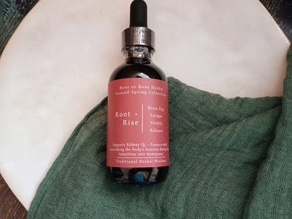 A glass dropper bottle of Root + Rise herbal tincture with a dusty rose label, resting on a textured green cloth over a white ceramic dish.
