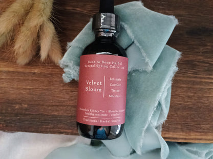 A dark amber glass bottle of Velvet Bloom herbal tincture with a dusty rose label, resting on a rustic wooden surface wrapped in a light blue cloth.