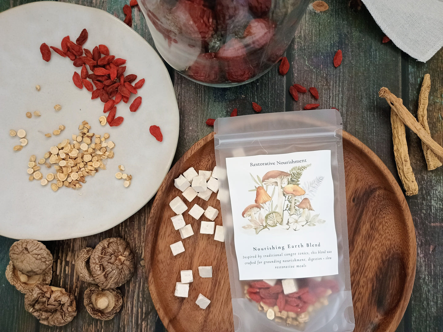 A clear stand-up pouch labeled 'Nourishing Earth Blend' Restorative Nourishment, filled with dried red and beige ingredients, arranged on a wooden tray with loose herbs, mushrooms, and seeds on a rustic dark wood surface.
