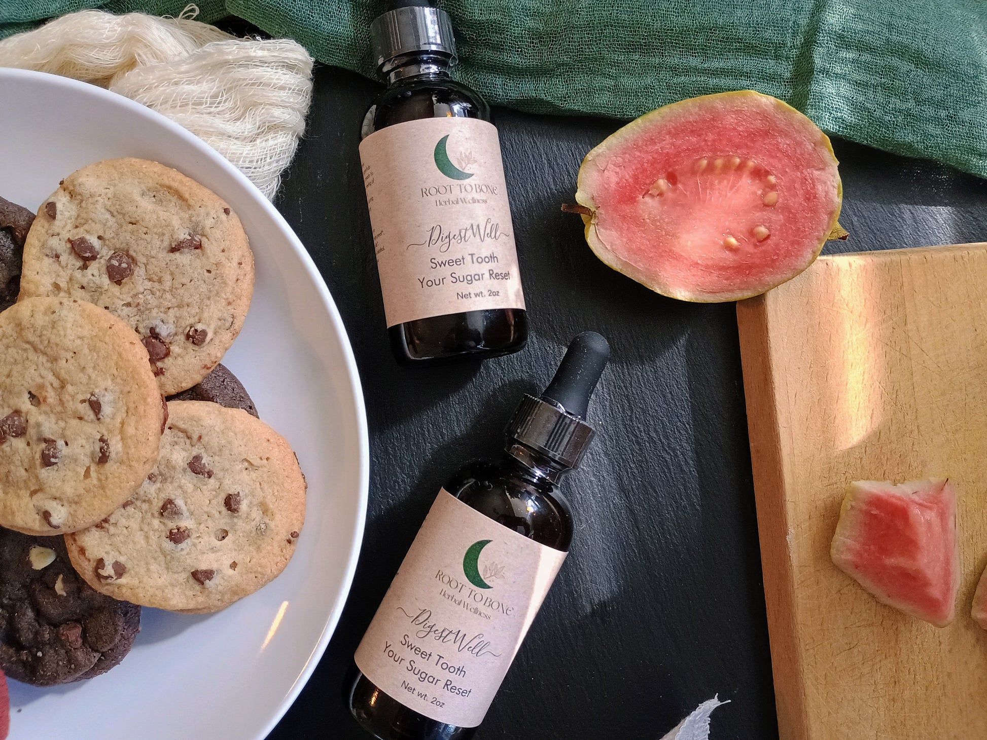 Two bottles of 'Root to Bone herbal' tincture on a dark surface with cookies and a watermelon slice.
