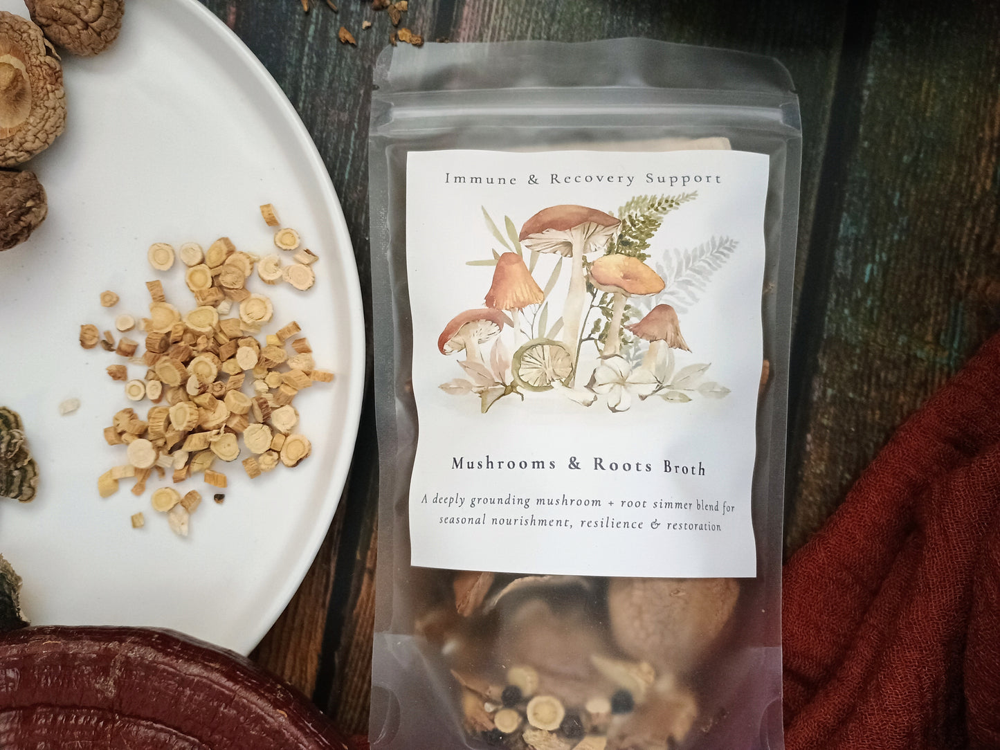 A clear stand-up pouch labeled 'Immune & Recovery Support' and 'Mushrooms & Roots Broth' sits on a dark wooden surface beside a white plate with sliced herbs and dried mushrooms. The pouch contains a brown herbal liquid, and a rust-red cloth is visible at the lower right.