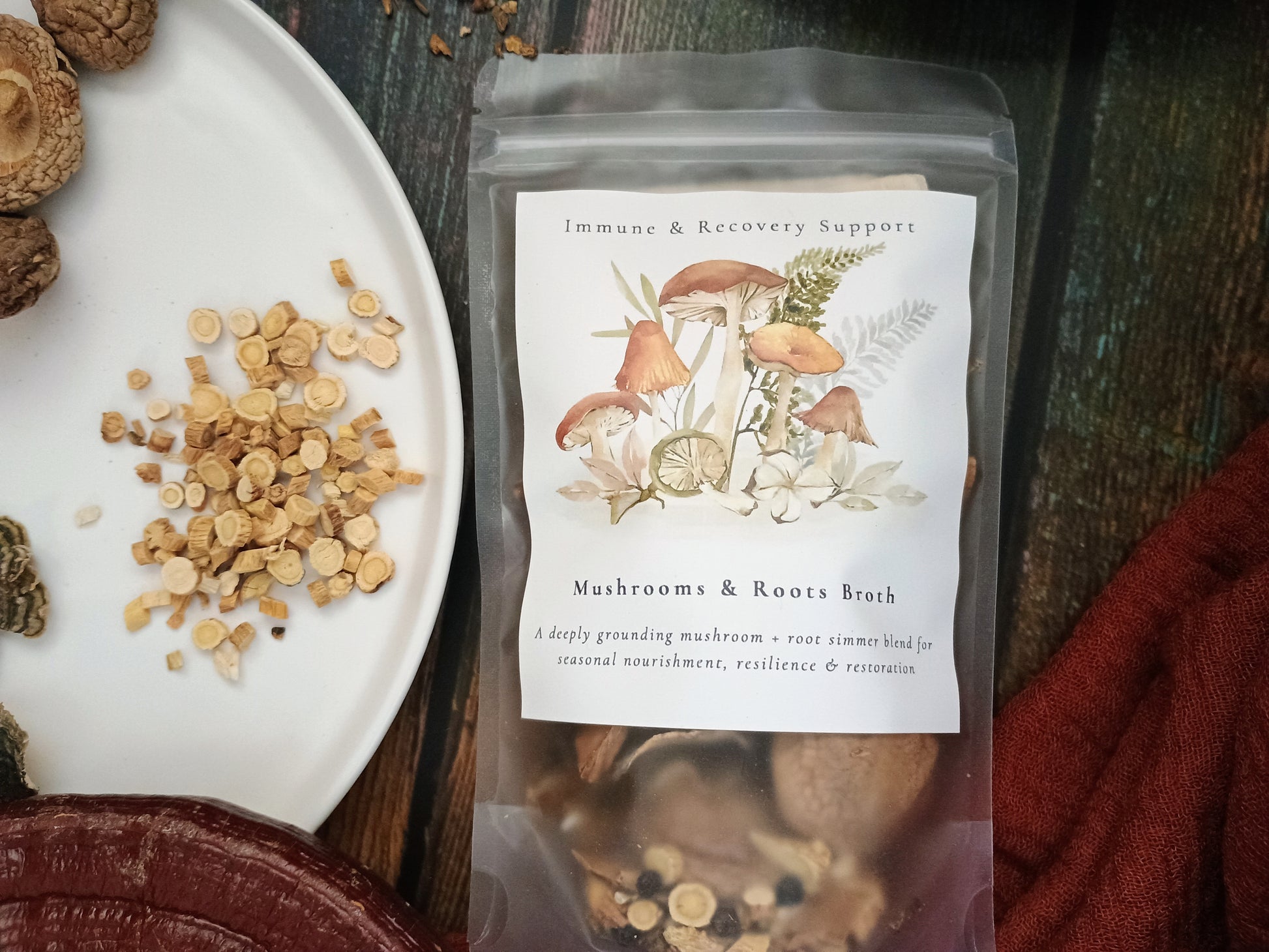 A clear stand-up pouch labeled 'Immune & Recovery Support' and 'Mushrooms & Roots Broth' sits on a dark wooden surface beside a white plate with sliced herbs and dried mushrooms. The pouch contains a brown herbal liquid, and a rust-red cloth is visible at the lower right.