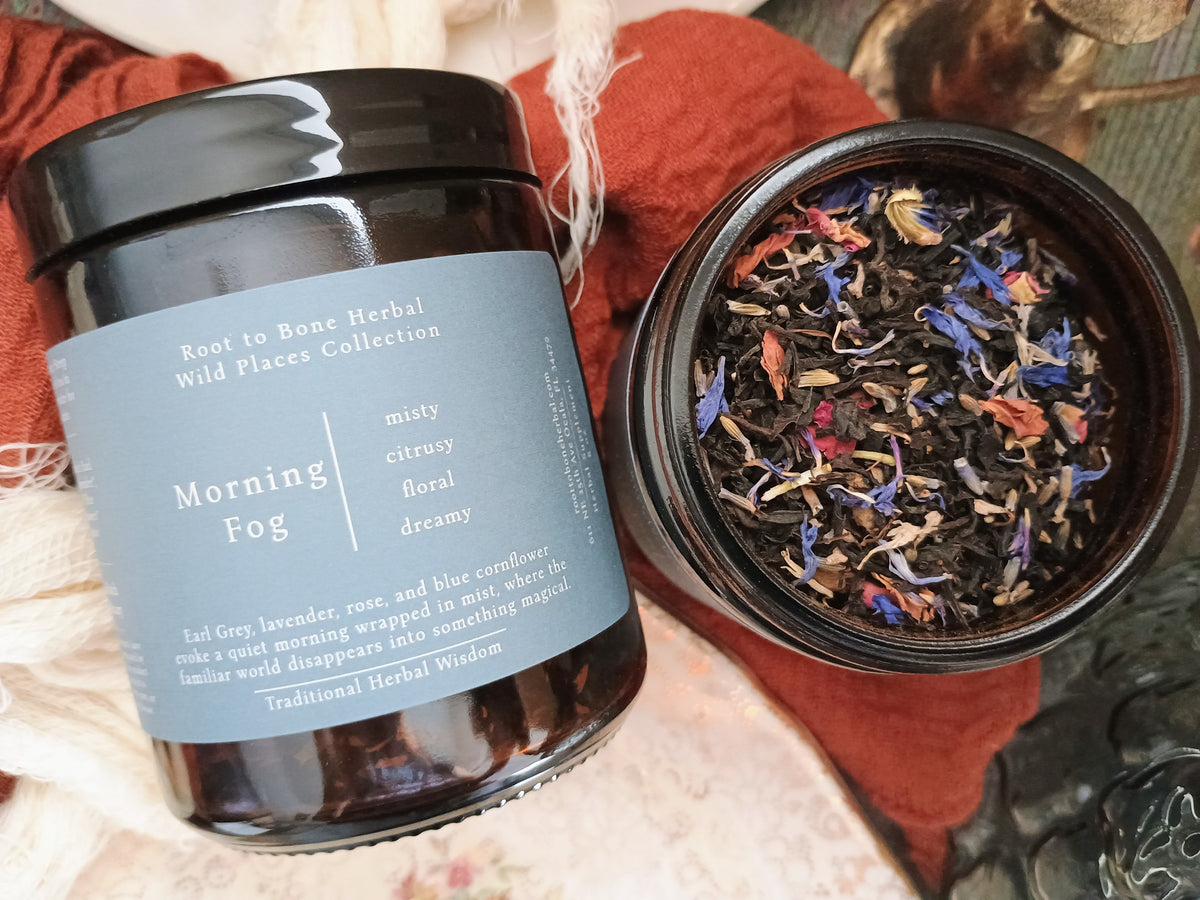 An amber glass jar candle labeled 'Morning Fog' from the Root to Bone Herbal Wild Places Collection, shown next to an open jar displaying the herbal blend inside. The candle is styled on a rust-colored fabric with soft cream-colored yarn.