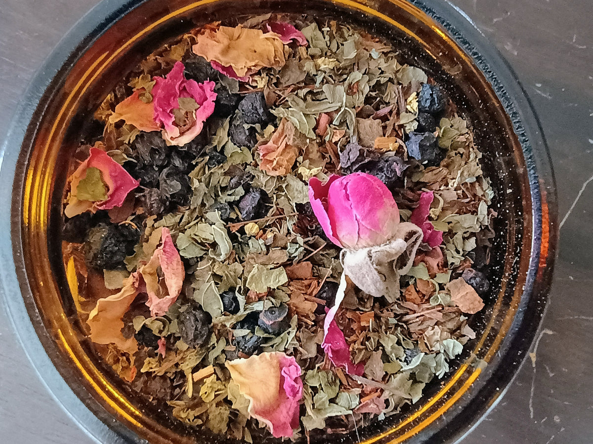 A top-down view of a round amber glass bowl filled with loose-leaf herbal tea, featuring dried green leaves, pink rose petals, and dark berries on a dark gray marble surface.