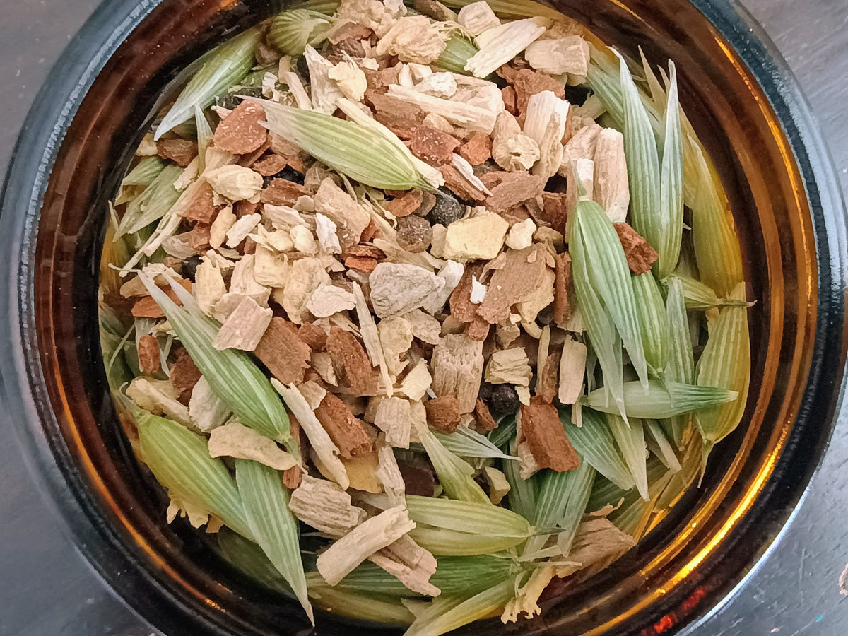 A top-down view of dried herbal bark pieces and green leaves inside a dark amber glass jar.