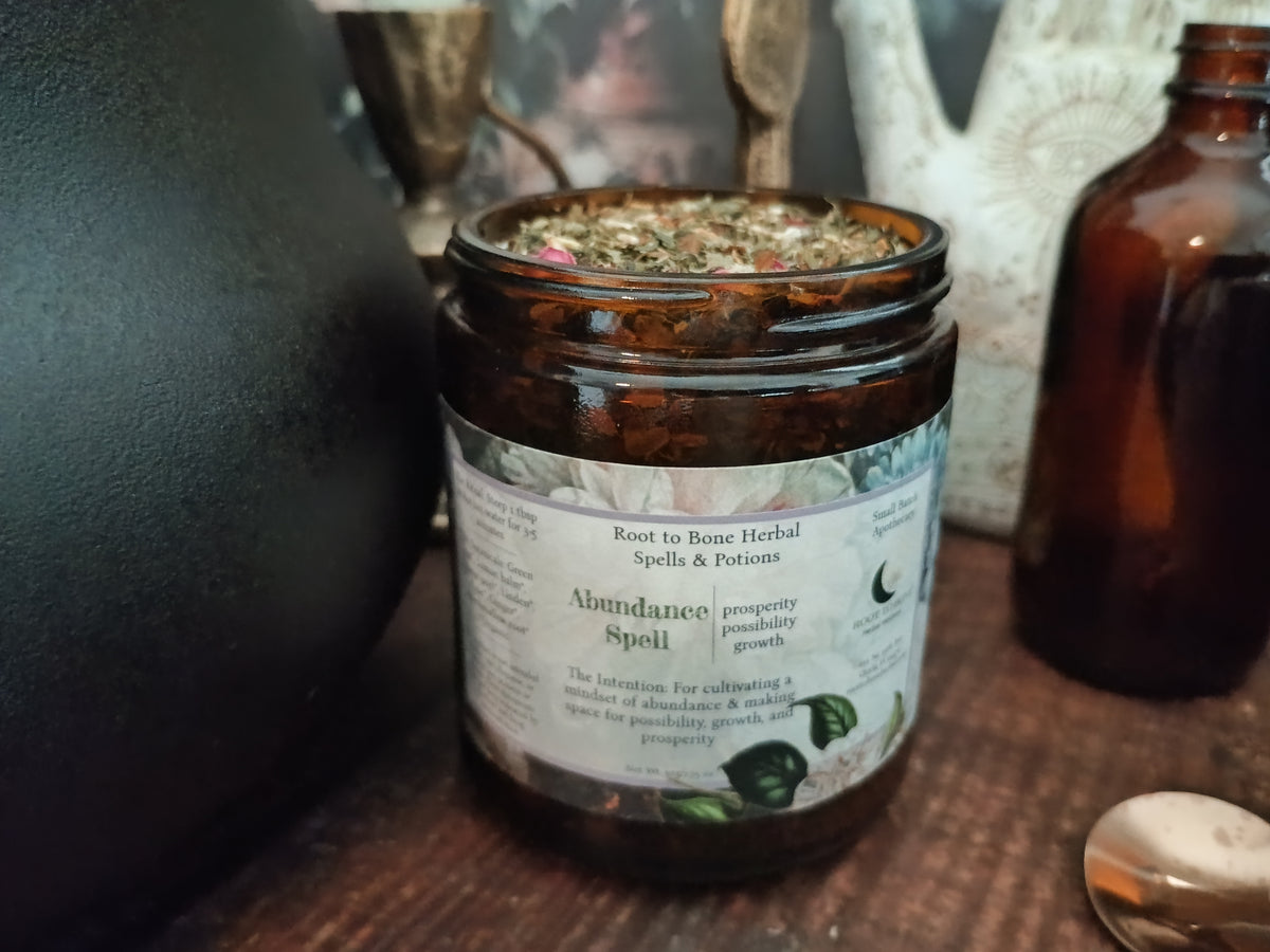 An amber glass jar candle labeled 'Abundance Spelt' by Root to Bone Herbal Spells & Potions, filled with dried herbs, set on a wooden surface next to an amber bottle.