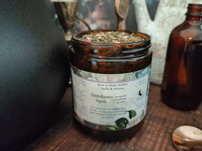 An amber glass jar candle labeled 'Abundance Spelt' by Root to Bone Herbal Spells & Potions, filled with dried herbs, set on a wooden surface next to an amber bottle.