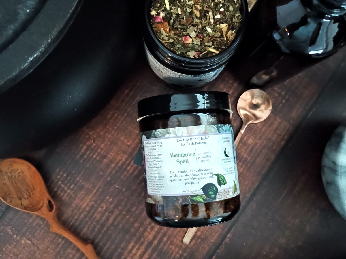 A dark amber glass jar of 'Abundance Spell' by Roost to Bone Herbal, styled on a dark wooden table with copper spoons and a pot of dried herbs.