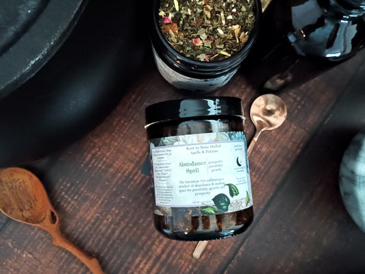 A dark amber glass jar of 'Abundance Spell' by Roost to Bone Herbal, styled on a dark wooden table with copper spoons and a pot of dried herbs.