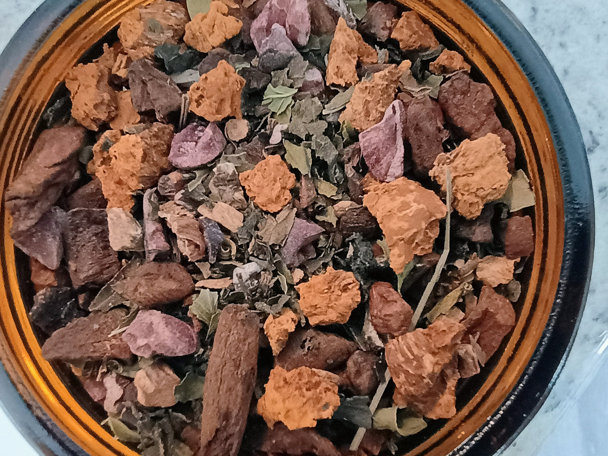 A close-up top-down view of loose herbal tea ingredients inside a dark amber glass bowl, including orange-brown bark pieces, dried leaves, and small purple flowers.