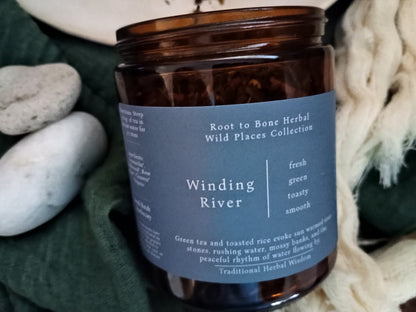 An amber glass jar with a slate blue label reading 'Winding River' and 'Root to Bone Herbal Wild Places Collection,' styled with dark green fabric, smooth stones, and cream-colored textiles.