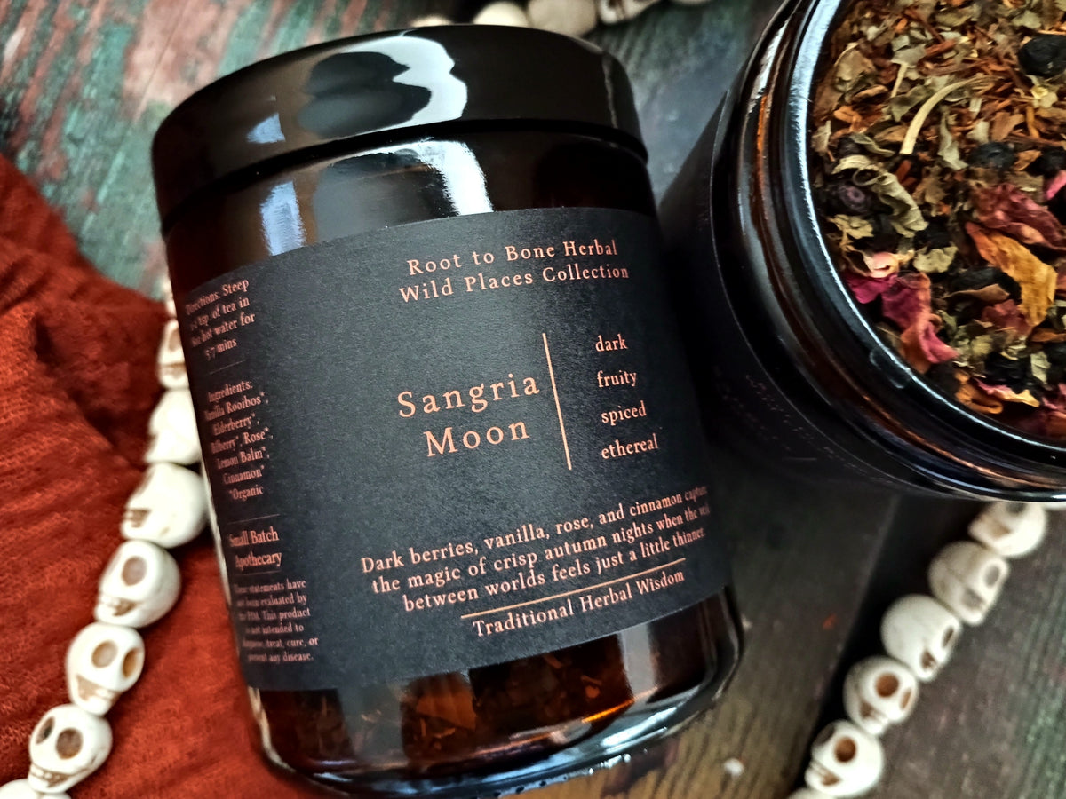 A dark amber glass jar candle labeled 'Sangria Moon' from the 'Root to Bone Herbal Wild Places Collection' sits on a rustic wooden surface next to an open jar showing dried herbs and flowers. Decorative white beads and a rust-colored fabric are scattered around.