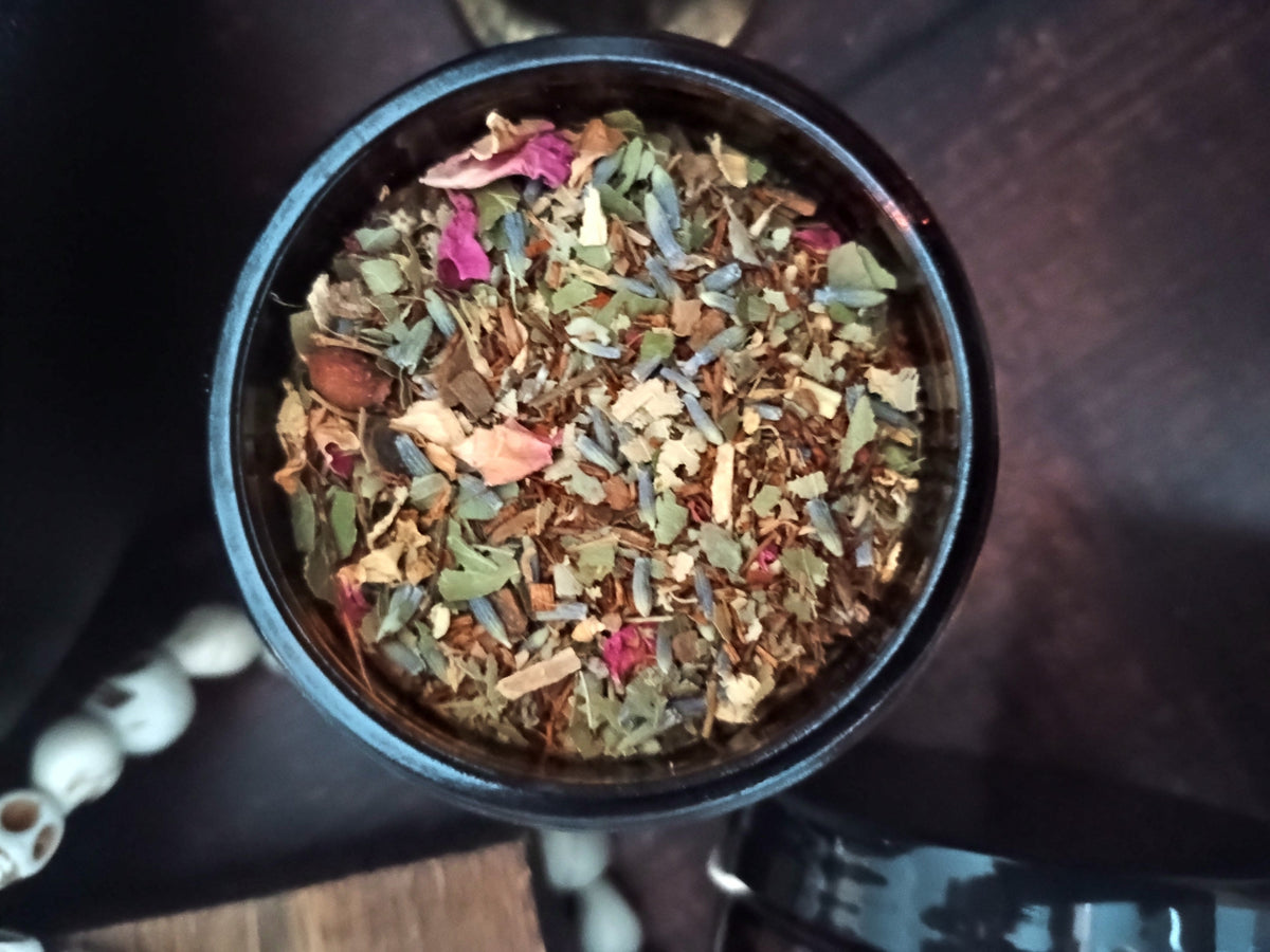 A top-down view of a black round tin filled with loose-leaf herbal tea blend, featuring dried flowers, leaves, and botanicals, set on a dark wooden surface.