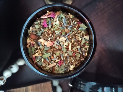 A top-down view of a black round tin filled with loose-leaf herbal tea blend, featuring dried flowers, leaves, and botanicals, set on a dark wooden surface.