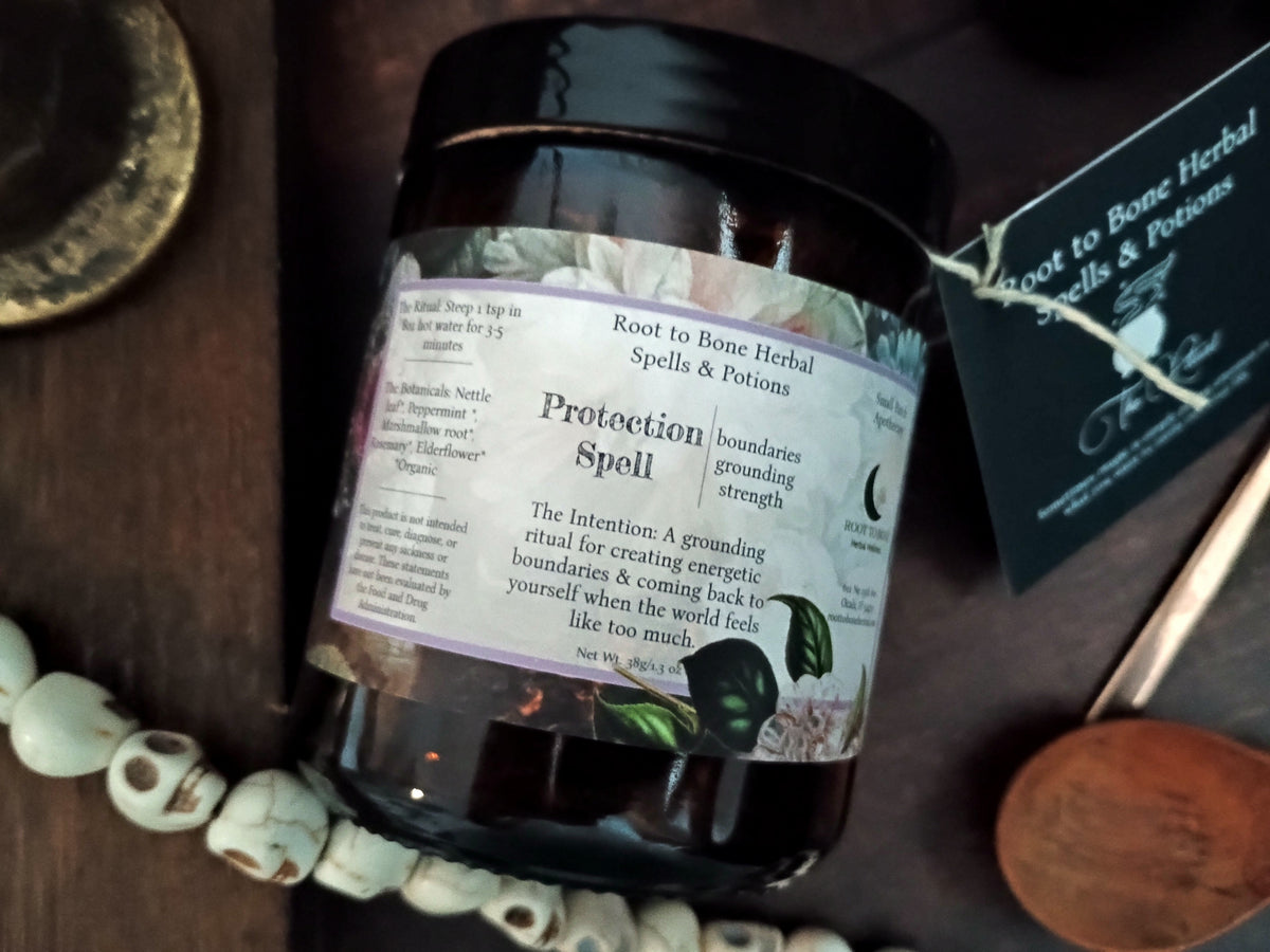 A dark glass jar labeled 'Protection Spell' by Root to Bone Herbal Spells & Potions, styled on a dark wooden surface with a wooden spoon, white beads, and a branded card.
